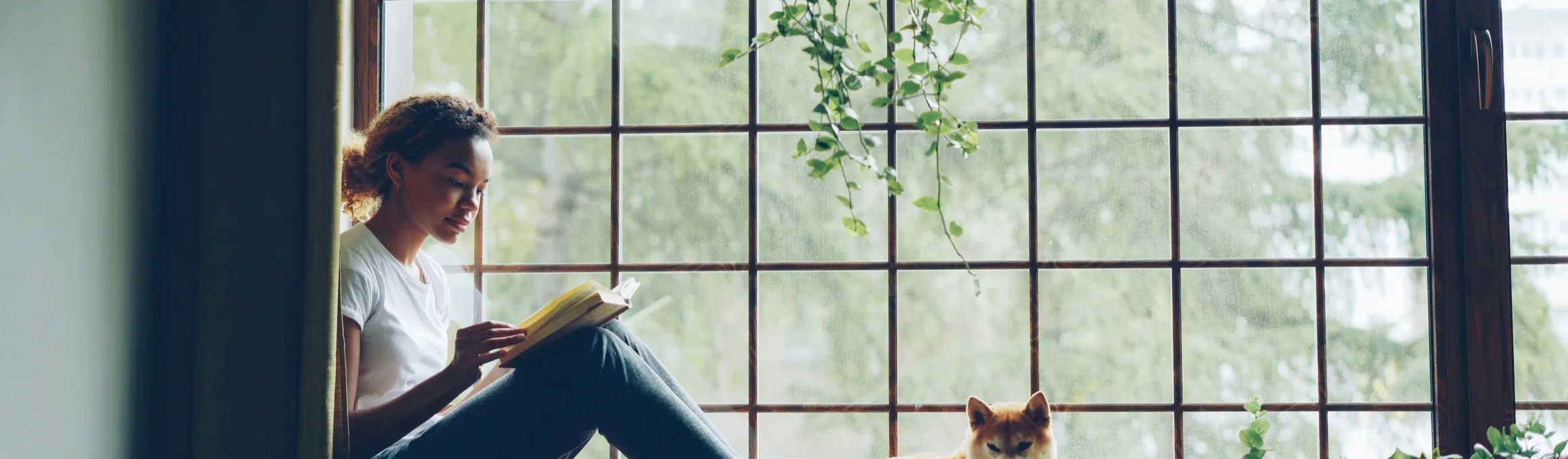 A woman reading by a sunlit window at home, her dog resting beside her